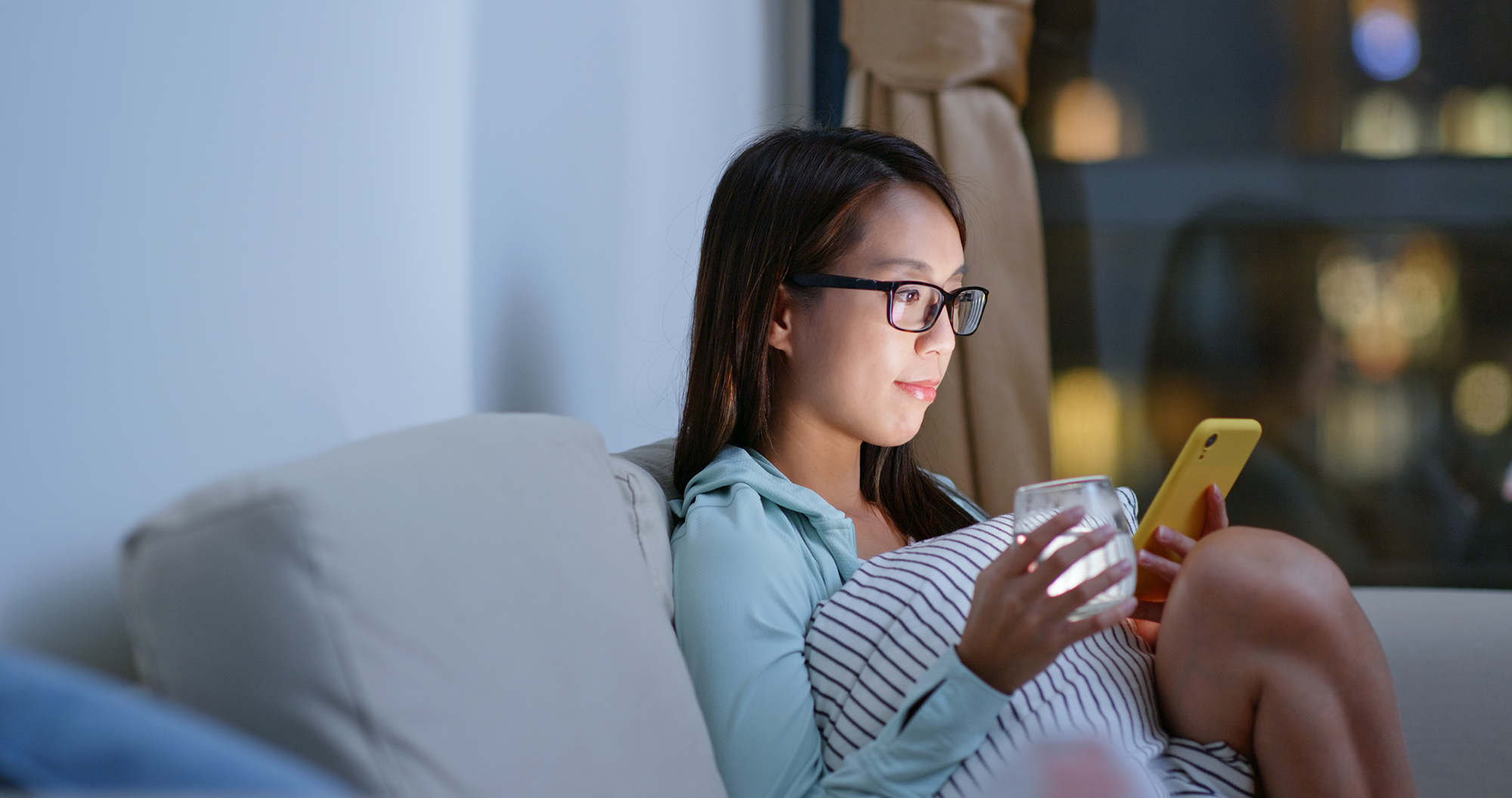 An Asian woman with dark hair and glasses sitting on the couch at night holding a glass of water and looking at her phone.