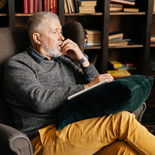 A senior is sitting in an armchair in his study. There is a pillow with a jounal on top of it on his lap. He is gazing into the distance with a concerned look.