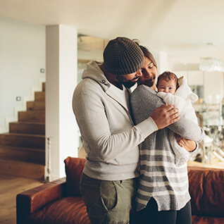 A mixed race couple is standing in their living room. The mother holds a new born baby, the father is embracing them from behind.