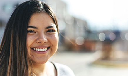 Portrait shot of smiling woman's face. She is standing in fron of a blurred background of some buildings on a sunny day.