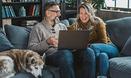 Man, woman, and dog sitting on a couch in their living room while the man and woman are laughing and looking at a laptop.