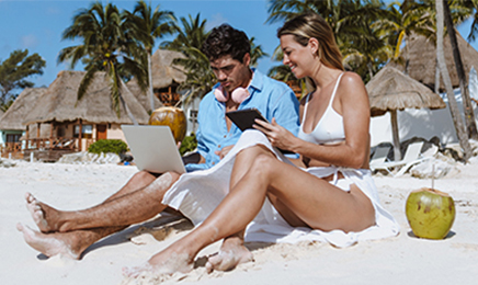 Couple sitting on beach while looking at electronic devices.