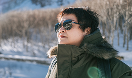 Person in green winter jacket wearing sunglasses in front of a snowy forest on a sunny day.