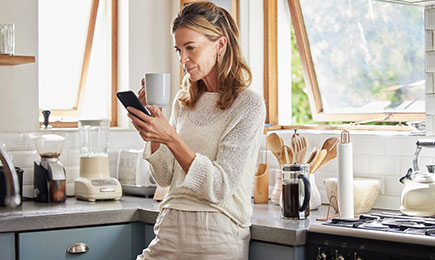 A woman is leaning against her kitchen counter while she drinks coffee, looking at her phone.